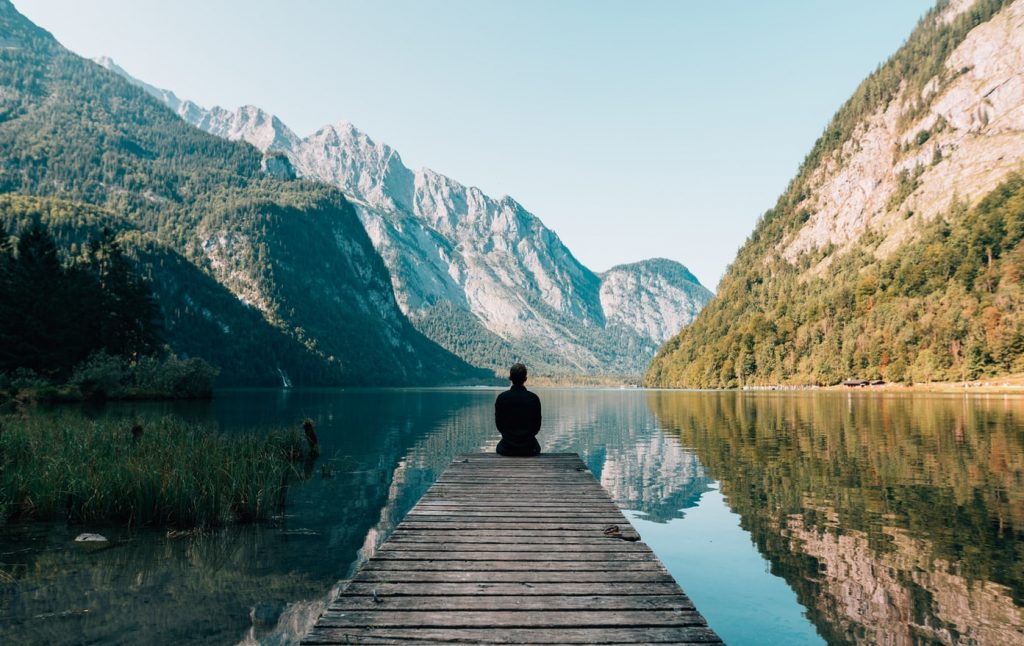 A person sitting on the end of a small pier, surrounded by a lake between two mountains